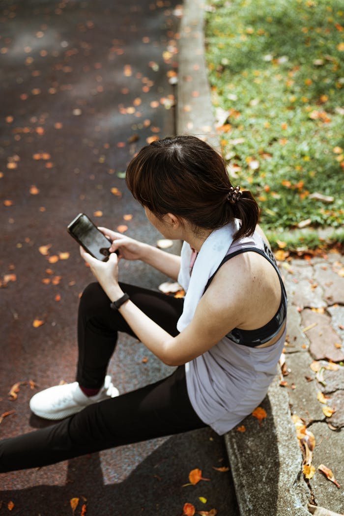 From above of female wearing sport clothes sitting on border and browsing smartphone while resting and searching information in internet